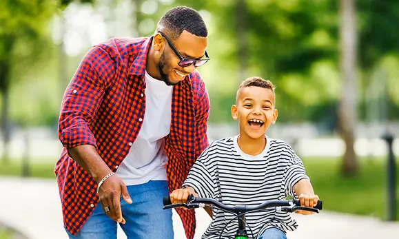 Padre ense&ntilde;ando a su hijo a manejar bicicleta felices en referencia a la P&oacute;liza de Seguro de Accidentes Personales Individual de Seguros Caracas